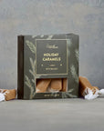A box of Ground Provisions Holiday Caramels with sea salt, displayed on a gray surface with two unwrapped caramels and white wrapping paper in the foreground.