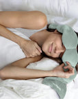 Person lying in bed wearing a green sleep mask with a white pillow and blanket.