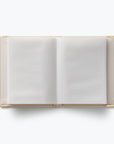 An open hardcover notebook with blank white pages and a light beige cover, framed by gold-toned metal corner protectors, shot from a top-down perspective on a white background.