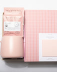 A pink gift set featuring a matte blush pink ceramic mug, a pink tea packet labeled 'TEA WITH TAI', a pink-and-white checkered notebook, and a white card labeled 'STICKY NOTES' on a white background.