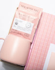 A pink ceramic mug sits next to a pink notebook with a grid pattern, a pink tea packet labeled 'TEA WITH TAE', and a sheet of sticky notes on a white background.