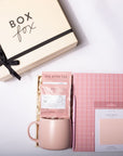 A styled gift set on a white background featuring a cream-colored 'Box Fox' gift box with a black ribbon, a pink ceramic mug, a pink tea packet labeled 'Tea with Tae', a pink grid notebook, and a pink sticky note pad.