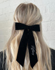 Person with long blonde hair wearing a black bow with 'Tiffany' branding against a white brick wall.