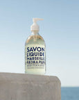 Bottle of Savon Liquide Marseille Extra Pur soap on a concrete ledge with a blue sky and ocean background.