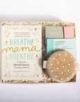 A curated gift box set for busy moms, featuring a book titled 'Breathe Mama Breathe' by Shonda Mensha, a peppermint eucalyptus diffuser, a pink bath bomb, and a natural cell resurfacer eye mask, all arranged in a kraft paper box with shredded paper filler on a white background.