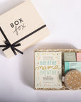 A gift set from Box Fox featuring a cream-colored box with a black ribbon, alongside an open box containing a 'Breathe Mama Breathe' mindfulness card, small bath or body items, and a round container of bath bombs, arranged on a white background.