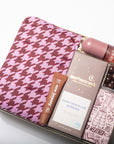 A curated gift box set on a white background, featuring a pink and burgundy houndstooth pouch, a box of Moonstruck Dark Chocolate Espresso, an Avery soap, and a Weatherproof pen in a clear plastic tray.
