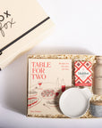 A curated gift box set from Box Fox, featuring the book 'Table for Two' by Bee Graham, a bottle of Le Marnier, a small white bowl, and a marble candle tin, arranged in a kraft paper box with shredded paper filler.