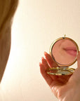 Hand holding a gold compact mirror against a plain background