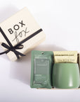 A gift set on a white background featuring a cream-colored 'BOX fox' gift box with a black ribbon, alongside an open package containing a sage green Noshinklu hand sanitizer packet and a matching sage green ceramic cup with two tea bags labeled 'TEA WITH TAE'.