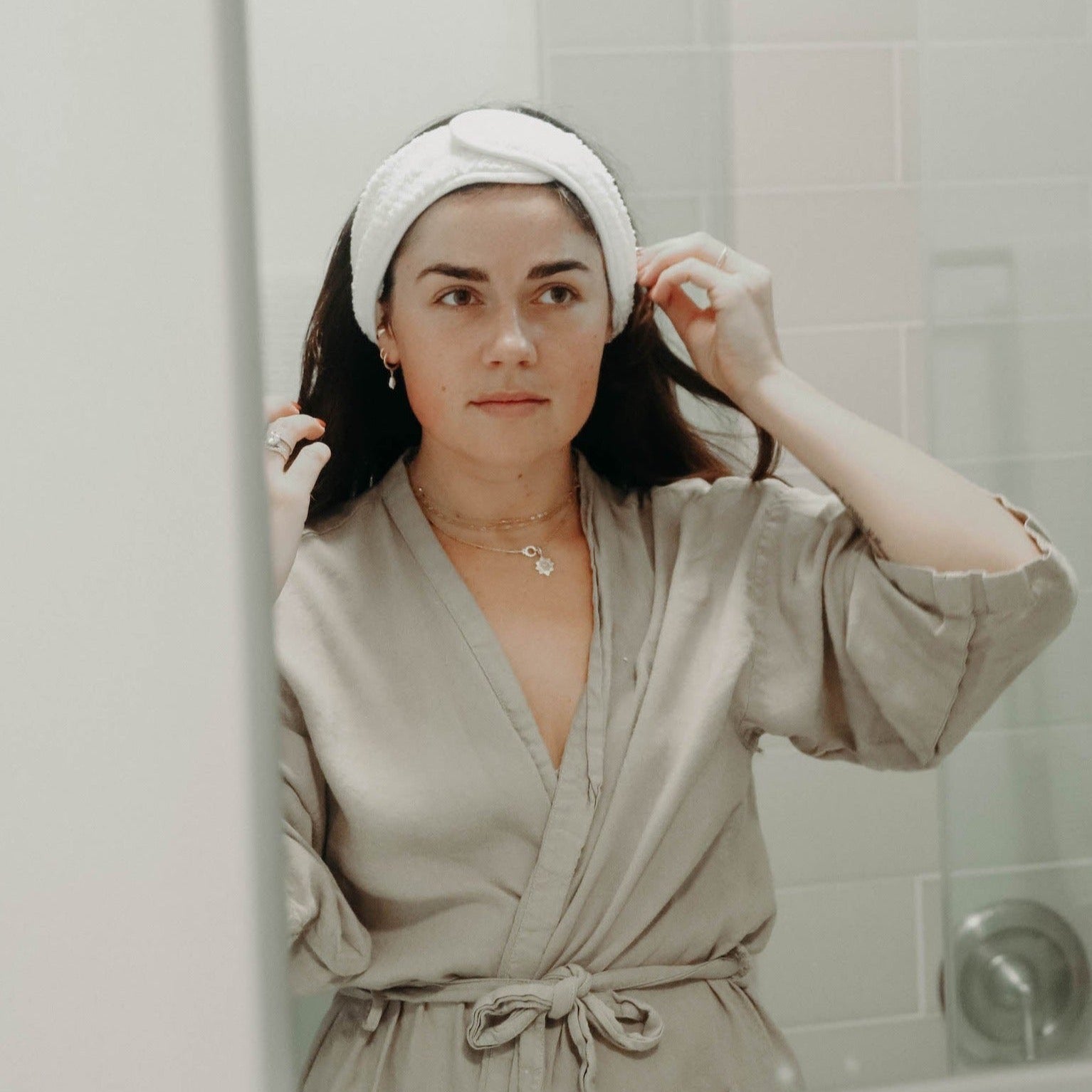 A woman with dark brown hair wearing a white plush spa headband and an oatmeal colored robe while she looks in the mirror.