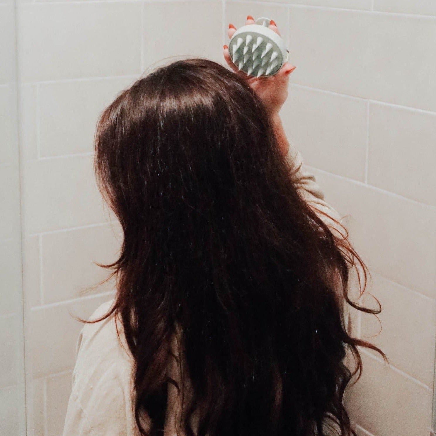 A woman with long wavy brown hair using a sage green scalp massager to massage the back of her head.