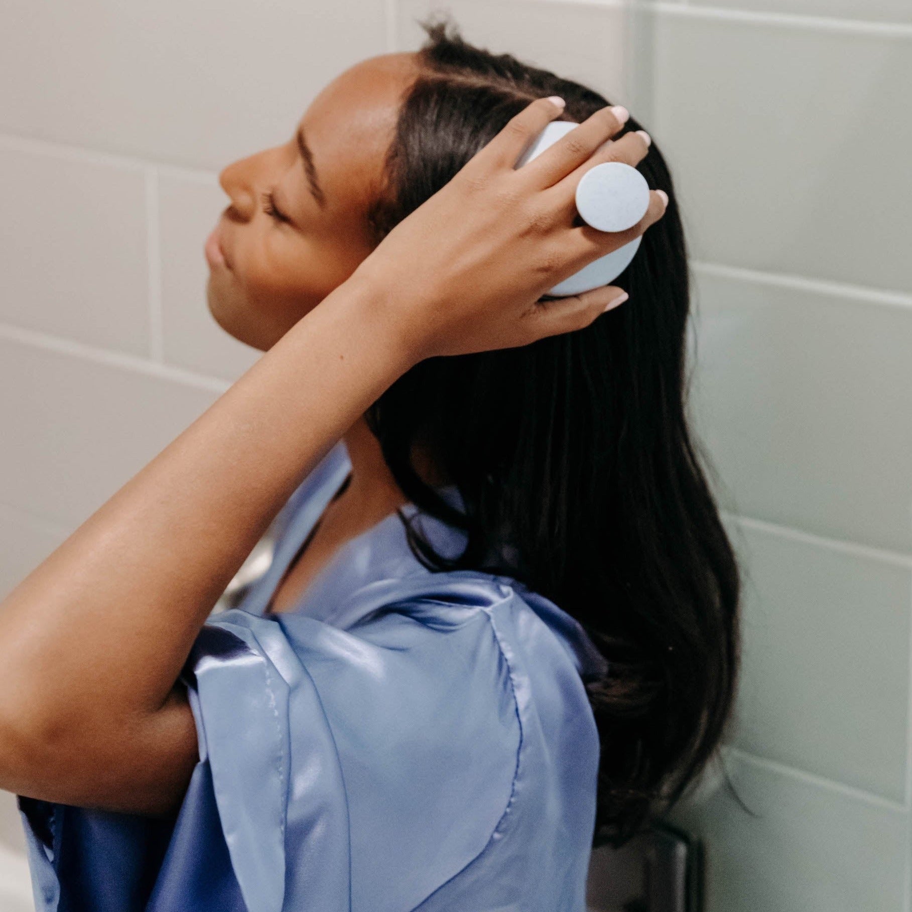 A girl with dark brown hair sitting in a shower using a mineral blue scalp massager on her scalp.