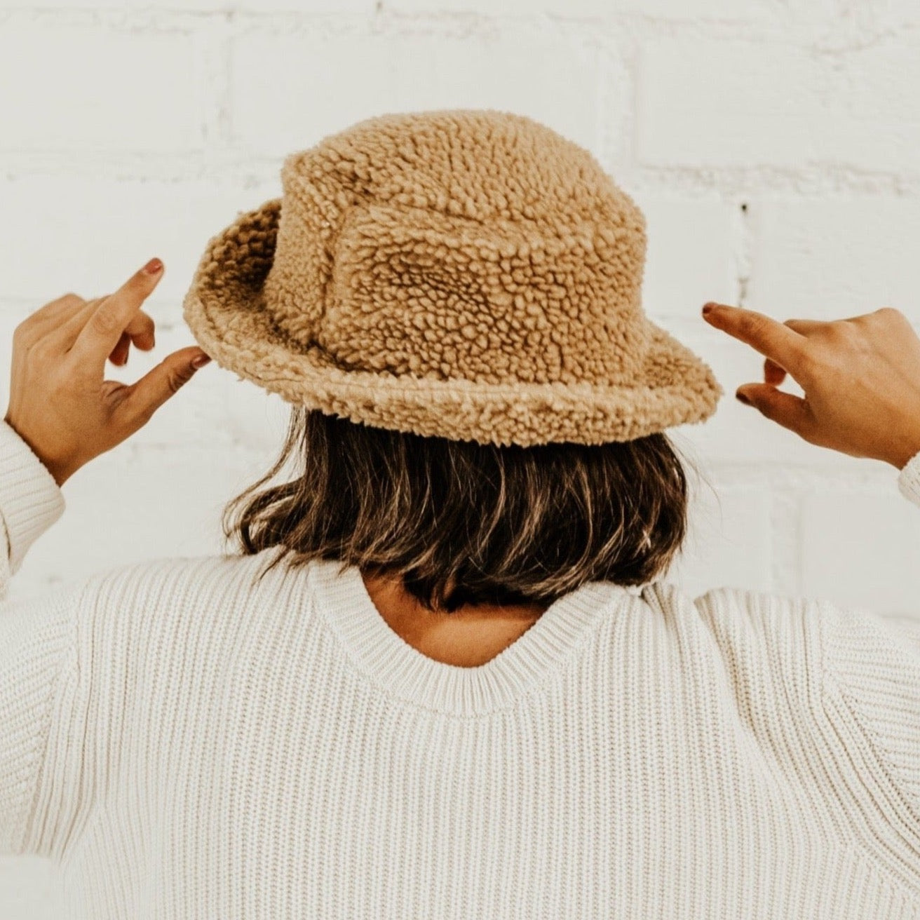A girl with short brown hair wearing a cream sweater and a brown sherpa teddy bucket hat. She is standing facing a white brick wall and the photo is taken from the waist up.