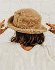 A girl with short brown hair wearing a cream sweater and a brown sherpa teddy bucket hat. She is standing facing a white brick wall and the photo is taken from the waist up.