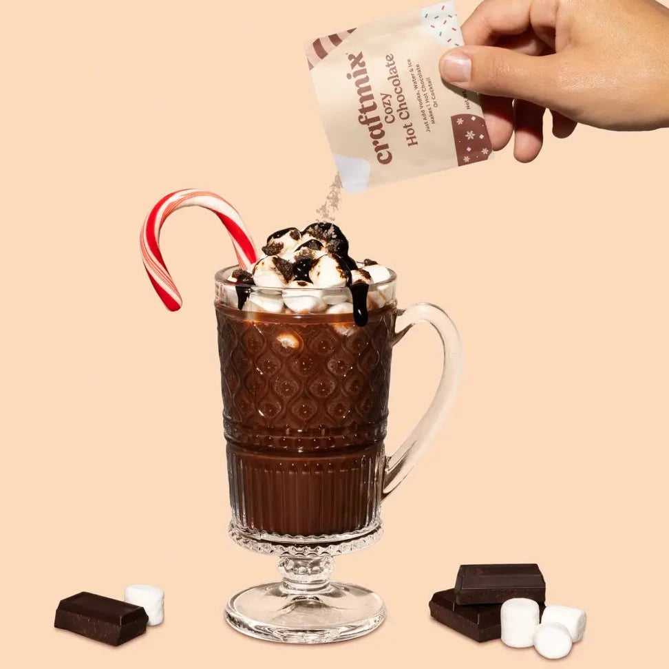 A glass mug of hot chocolate topped with whipped cream, marshmallows, and chocolate drizzle. A candy cane is placed inside the mug as garnish, with chocolate pieces and mini marshmallows scattered around. A hand is seen pouring in a packet of Craftmix Cozy Hot Chocolate mix