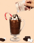 A glass mug of hot chocolate topped with whipped cream, marshmallows, and chocolate drizzle. A candy cane is placed inside the mug as garnish, with chocolate pieces and mini marshmallows scattered around. A hand is seen pouring in a packet of Craftmix Cozy Hot Chocolate mix