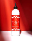 bottle of "Brightland Pizza Oil" set against a red background with lighting that creates a dramatic shadow effect. The bottle is white with a prominent red label featuring white checkered patterns on the top and bottom. The label reads "Brightland Pizza Oil," described as "Jalapeño & Herb Flavored Olive Oil," with a volume of "12 FL OZ (355 mL)." The bottle has a black nozzle cap designed for easy pouring.