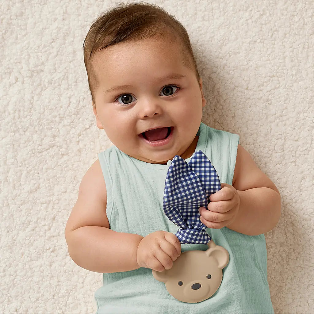 Baby holding a blue checkered bow tie and teddy bear toy against a beige background