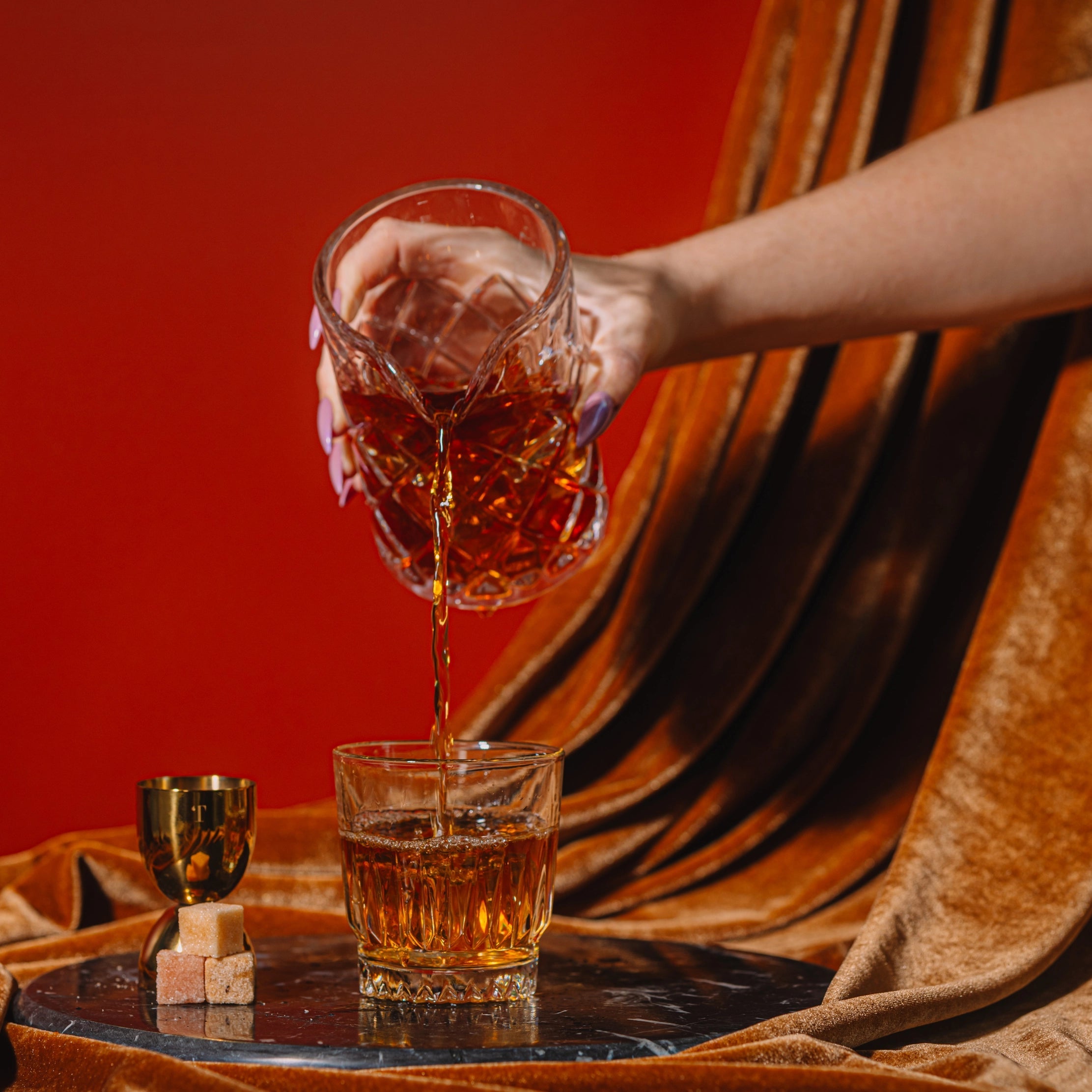 Hand pouring amber liquid from a glass into another glass on a draped fabric background