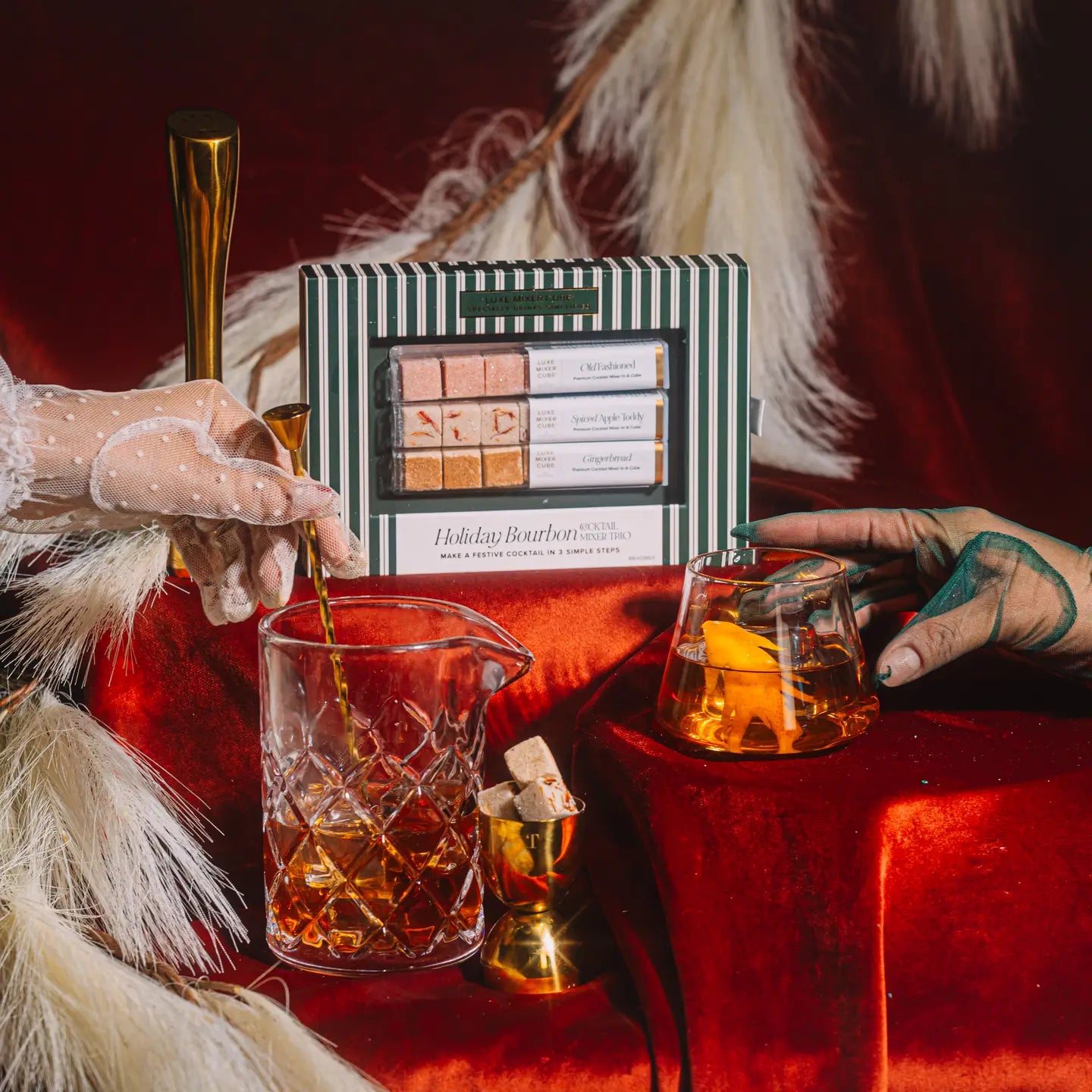 Two hands with gloves holding glasses of a drink on a red surface with a box of bourbon bars in the background.