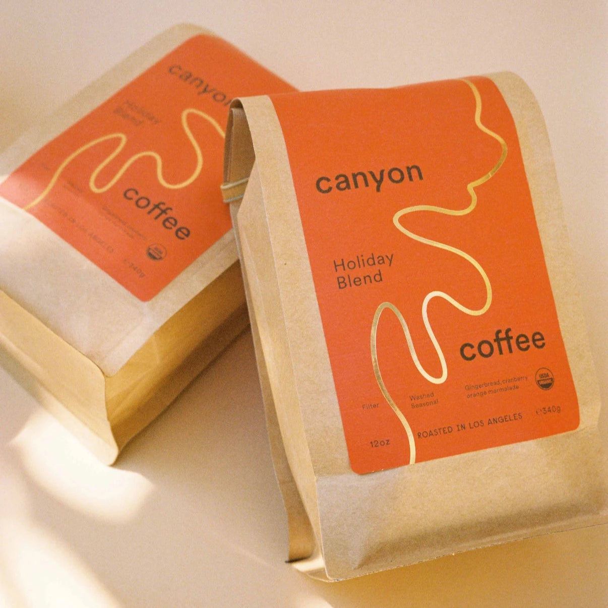 Two bags of Canyon Coffee's Holiday Blend lying on a soft-lit surface. The red and kraft paper packaging with gold accents is prominent, creating a warm and festive atmosphere.