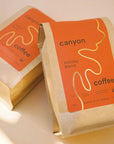 Two bags of Canyon Coffee's Holiday Blend lying on a soft-lit surface. The red and kraft paper packaging with gold accents is prominent, creating a warm and festive atmosphere.