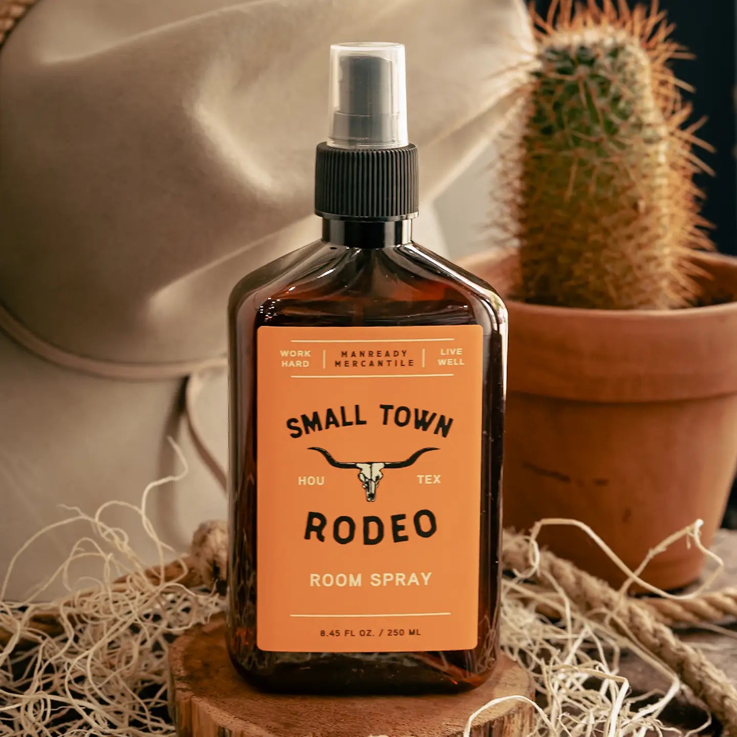 Room spray bottle labeled 'Small Town Rodeo' on a wooden block with a cactus in the background.
