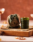 A product shot featuring a green votive candle labeled "Wine Under the Tree" with notes of blue spruce, cedar, and bright citrus. The candle is displayed next to its matching drawstring pouch on a rustic wooden tray, accompanied by star anise and dried citrus, evoking a warm, festive vibe.