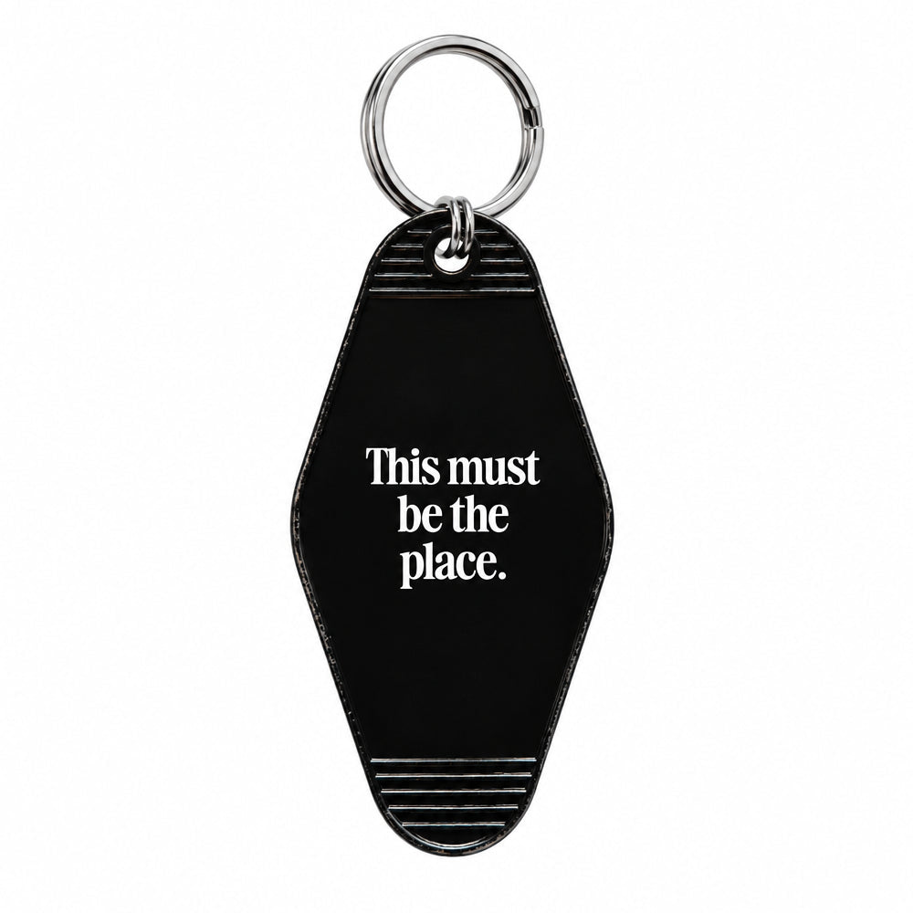 A black enamel pin shaped like a vintage motel room keychain, featuring the white text 'This must be the place.' and decorative silver striped details at the top and bottom, shown on a plain white background.