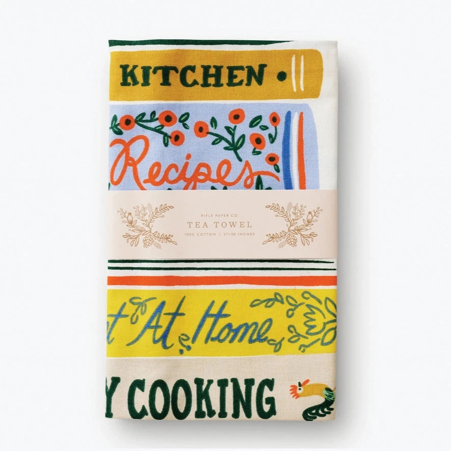 A folded tea towel showing a close-up of the same cookbook-themed design. The text "Kitchen" and "Recipes" is visible, along with floral decorations. A paper label wraps around the towel, reading "Rifle Paper Co. Tea Towel."