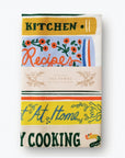 A folded tea towel showing a close-up of the same cookbook-themed design. The text "Kitchen" and "Recipes" is visible, along with floral decorations. A paper label wraps around the towel, reading "Rifle Paper Co. Tea Towel."