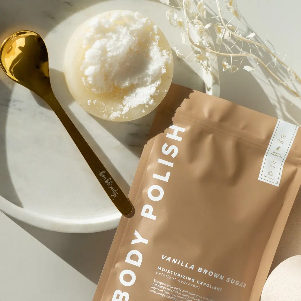 A stand-up pouch of body polish in Vanilla Brown Sugar moisturising exfoliant, styled on a white marble tray with a gold spoon and a round container of white exfoliating scrub.