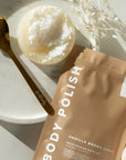 A stand-up pouch of body polish in Vanilla Brown Sugar moisturising exfoliant, styled on a white marble tray with a gold spoon and a round container of white exfoliating scrub.
