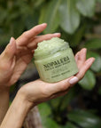 Hand holding a jar of Nopalera Cactus Body Polish against a green leafy background