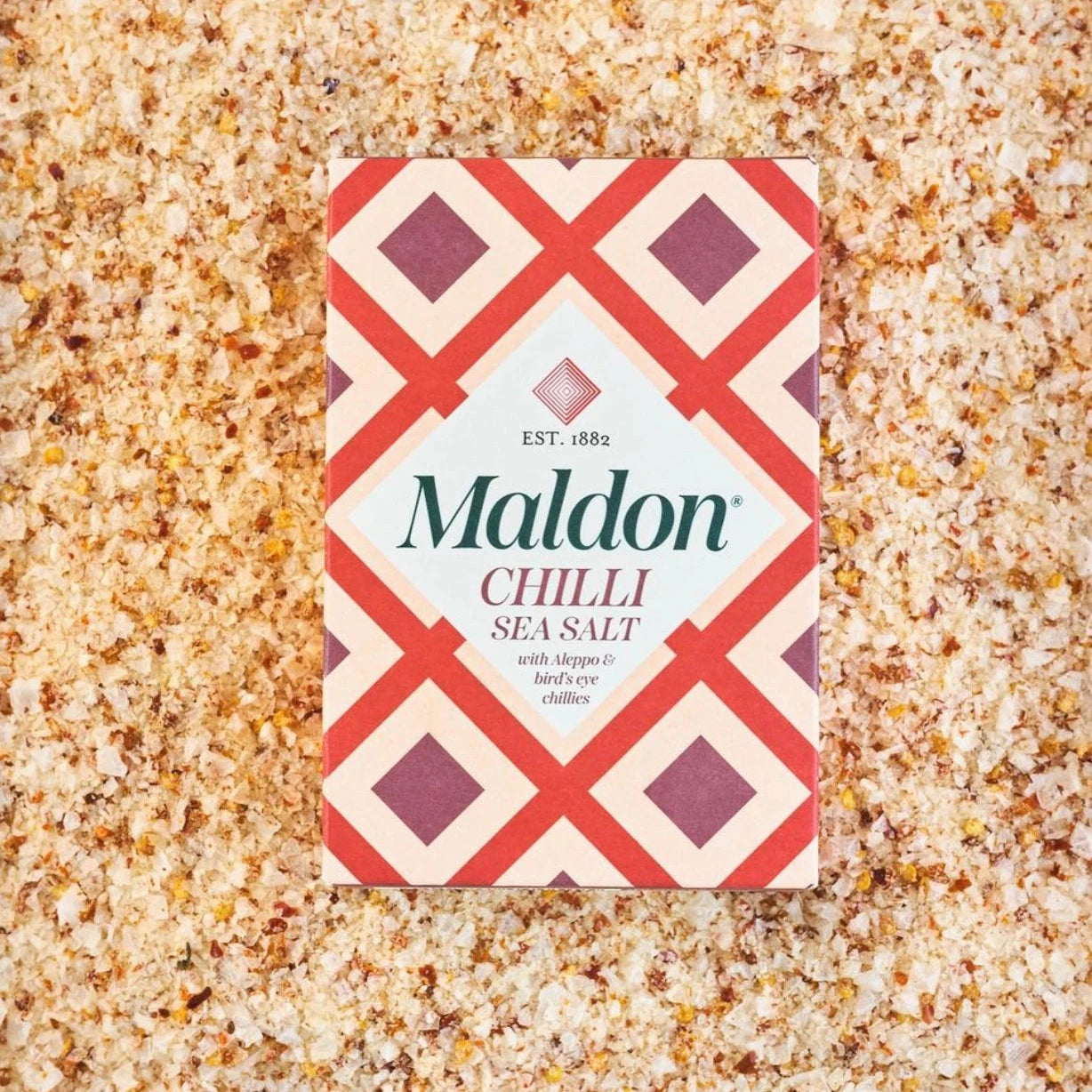 box of Maldon Chilli Sea Salt placed on a bed of the salt itself. The salt is mixed with red chili flakes, giving it a textured and vibrant appearance. The box has a red and cream-colored geometric design with the text "Maldon Chilli Sea Salt" and notes that it includes Aleppo and bird's eye chilies.
