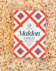 box of Maldon Chilli Sea Salt placed on a bed of the salt itself. The salt is mixed with red chili flakes, giving it a textured and vibrant appearance. The box has a red and cream-colored geometric design with the text "Maldon Chilli Sea Salt" and notes that it includes Aleppo and bird's eye chilies.
