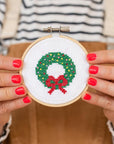 This image shows a small embroidery hoop held by someone with bright red nail polish. The cross-stitch design within the hoop features a festive wreath made up of green leaves and dotted with red, yellow, and green accents, mimicking holiday ornaments. At the bottom of the wreath is a red bow, adding to the seasonal theme. The person holding the embroidery is dressed casually in a striped shirt and overalls, suggesting a fun, homemade craft project. The image captures a warm, creative holiday spirit.