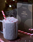 This image beautifully ties together the peppermint hot cocoa from "Grand Provisions" with a cozy, festive setup. The hot cocoa is presented in a rustic grey mug with melted chocolate elegantly dripping down the side. It's topped with fluffy whipped cream and sprinkled with crushed candy cane pieces, adding visual texture and a festive vibe. A candy cane stick completes the presentation, both as a decorative element and an enhancer for peppermint flavor.