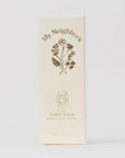 A tall cream-colored product box for 'My Neighbor's Hand Balm' featuring a dark brown botanical illustration and text, set against a clean, light gray background.
