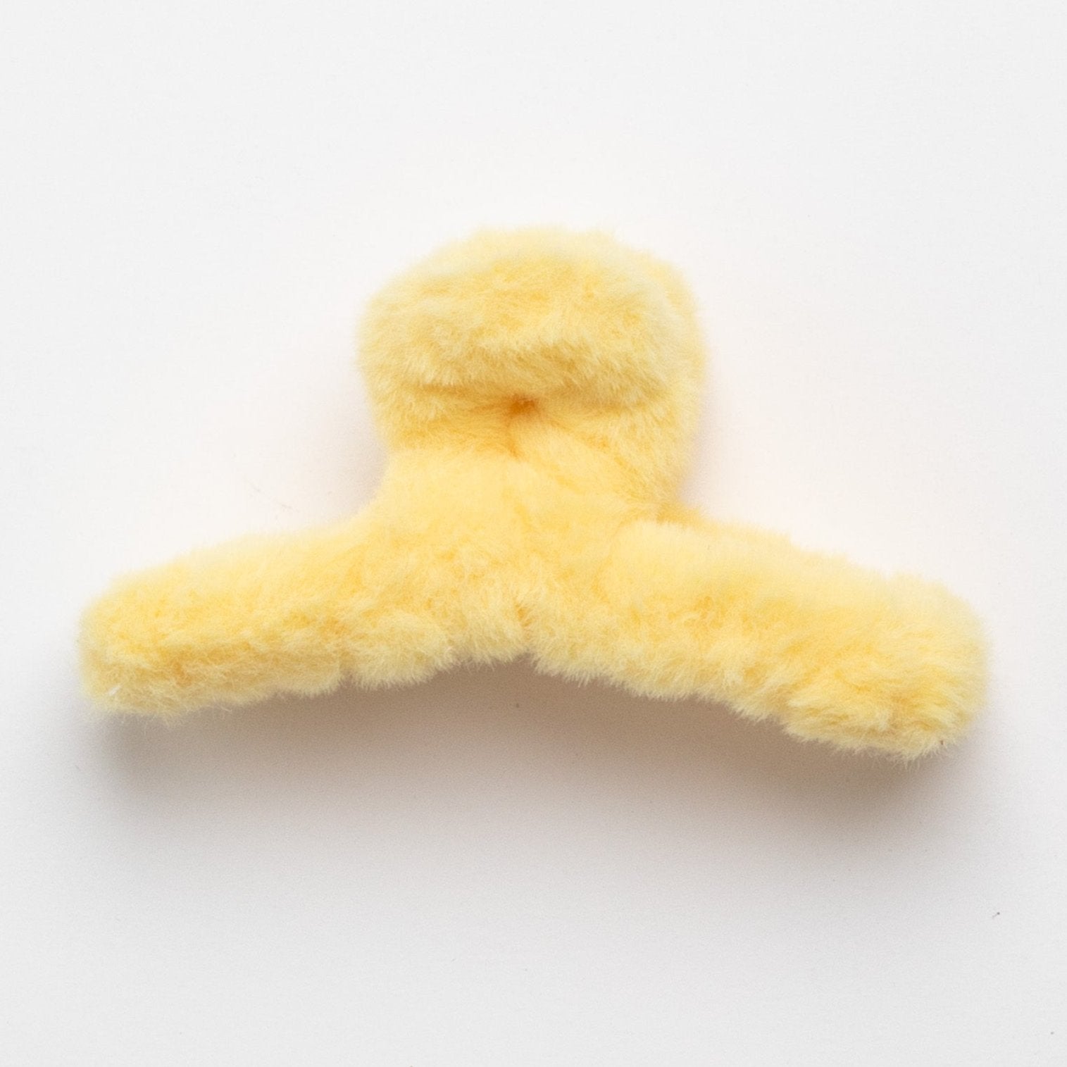 This image shows a soft, fluffy hair claw clip in a pale yellow color. The plush texture gives it a cozy appearance, and it is shaped with two gripping arms to hold hair securely. The clip is placed on a plain white background.