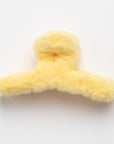 This image shows a soft, fluffy hair claw clip in a pale yellow color. The plush texture gives it a cozy appearance, and it is shaped with two gripping arms to hold hair securely. The clip is placed on a plain white background.