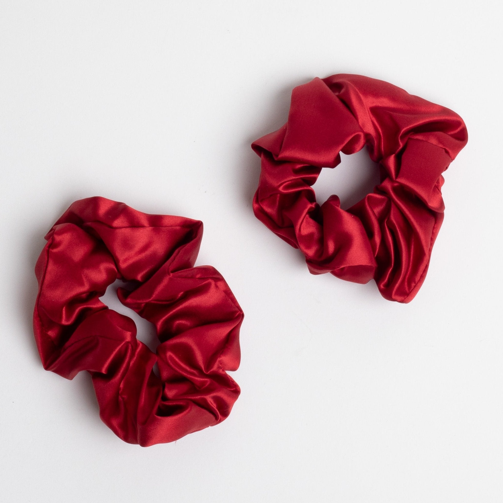 This image shows two red satin scrunchies, laid flat on a white background. The glossy fabric gives them a smooth and elegant appearance, ideal for tying hair without causing damage or creases.