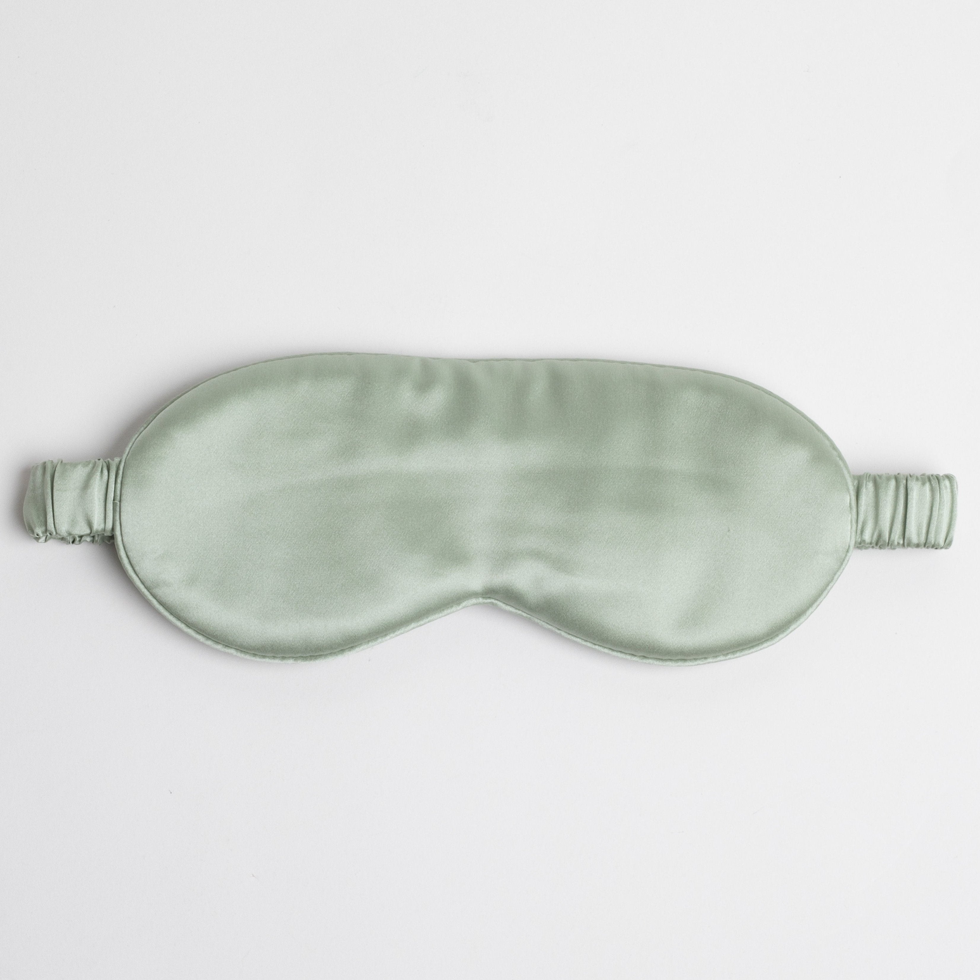 This image shows a silky light green sleep mask with an elastic strap. The smooth, glossy fabric suggests a comfortable feel, designed to block light and promote restful sleep. It is laid flat on a plain white background.The sleep mask in the image appears to be a soft sage green color. Its smooth, satin finish gives it a subtle sheen, enhancing the gentle green tone.
