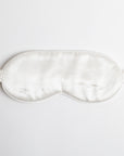 This image shows a silky white sleep mask with an elastic strap. The smooth, glossy fabric suggests a comfortable feel, designed to block light and promote restful sleep. It is laid flat on a plain white background.
