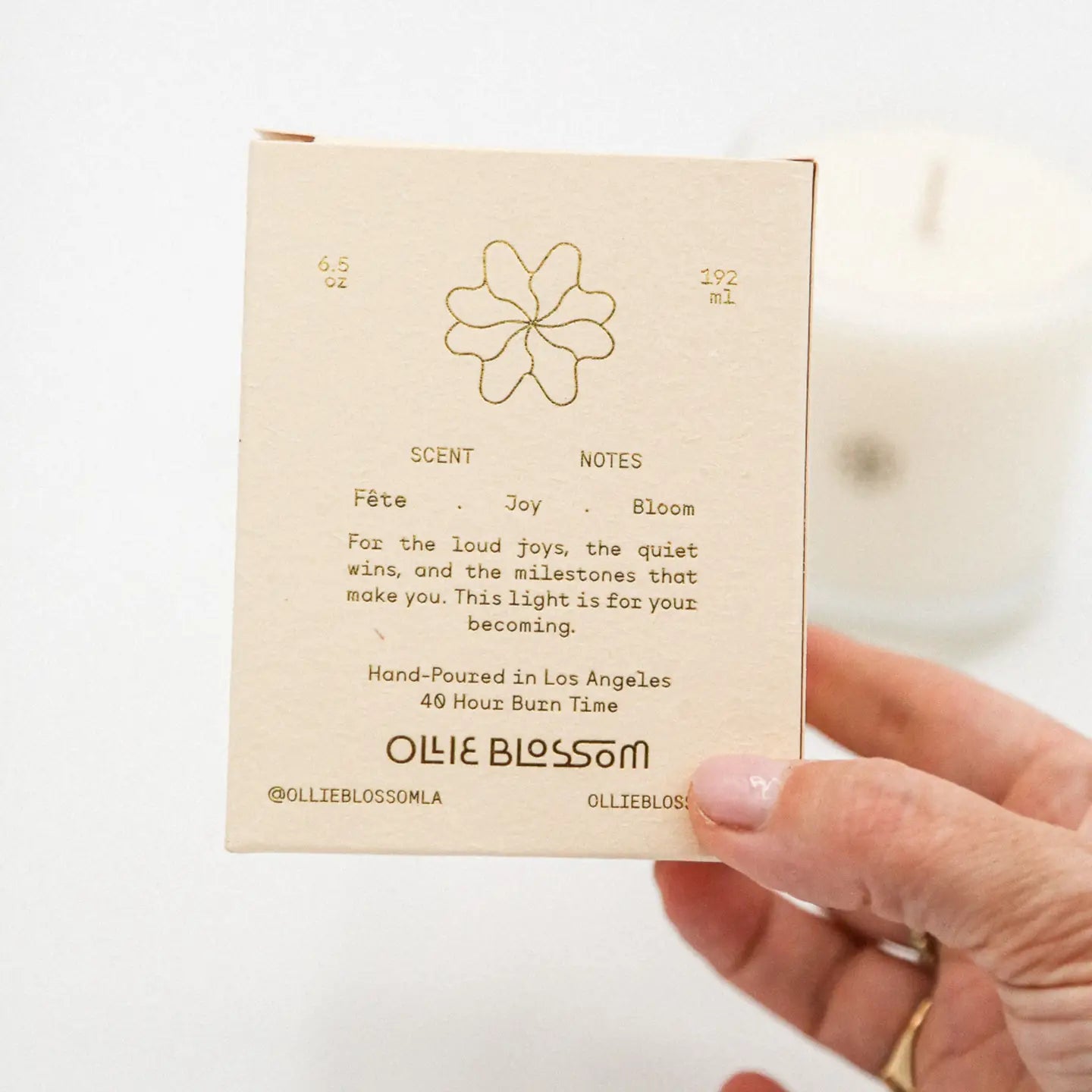 Hand holding a small beige box with text and a flower design, featuring the brand 'Ollie Blossom'.