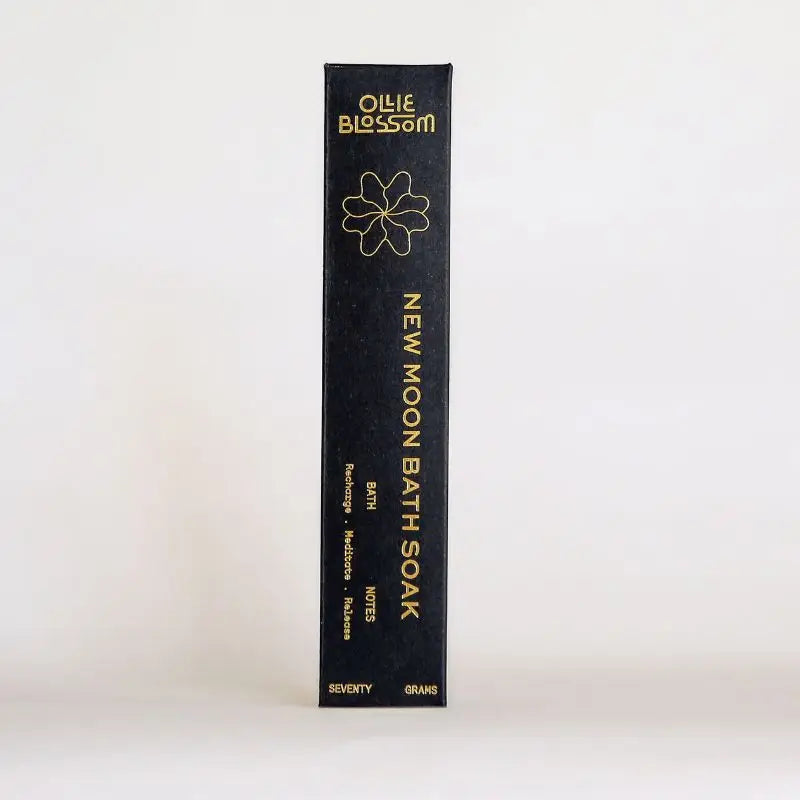 A tall, narrow black box for Ollie Blossom New Moon Bath Soak, featuring gold text and a gold outline flower logo, photographed against a clean white background.