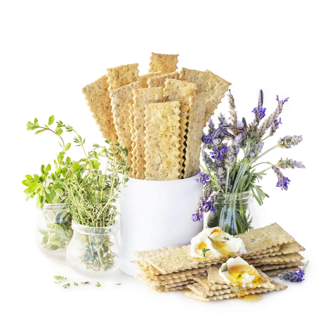 Flatbread crackers arranged in a white ceramic container with small glass jars of herbs and lavender on a white background.