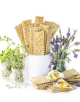 Flatbread crackers arranged in a white ceramic container with small glass jars of herbs and lavender on a white background.