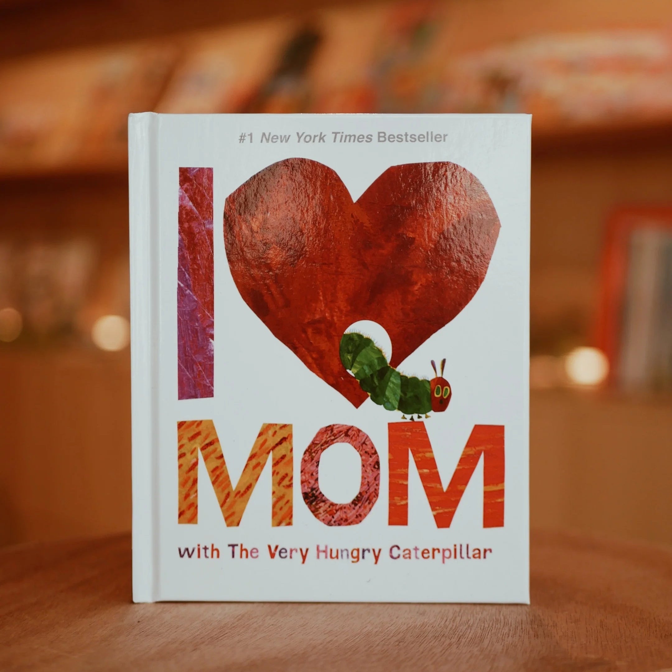 Book titled 'I Love Mom with The Very Hungry Caterpillar' on a wooden surface with a blurred bookshelf background.